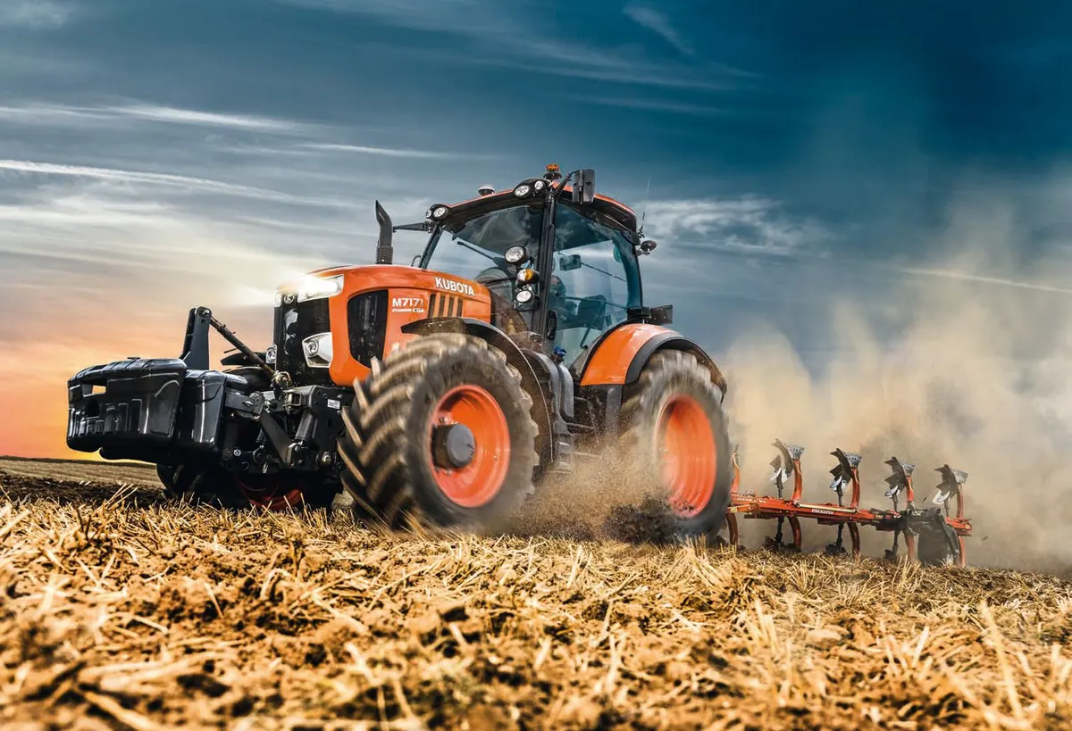 A Kubota M7 tractor ploughing a stubble field, dust trailing behind it